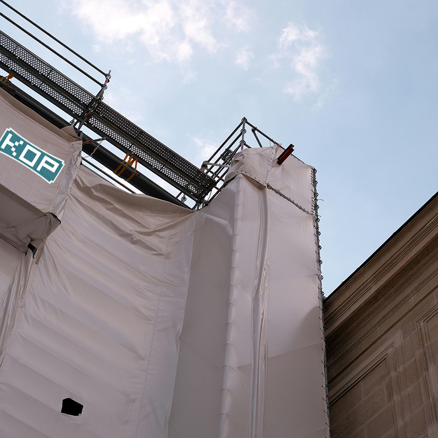 A building under construction is covered in white protective sheeting and scaffolding, with clear blue sky and clouds in the background.