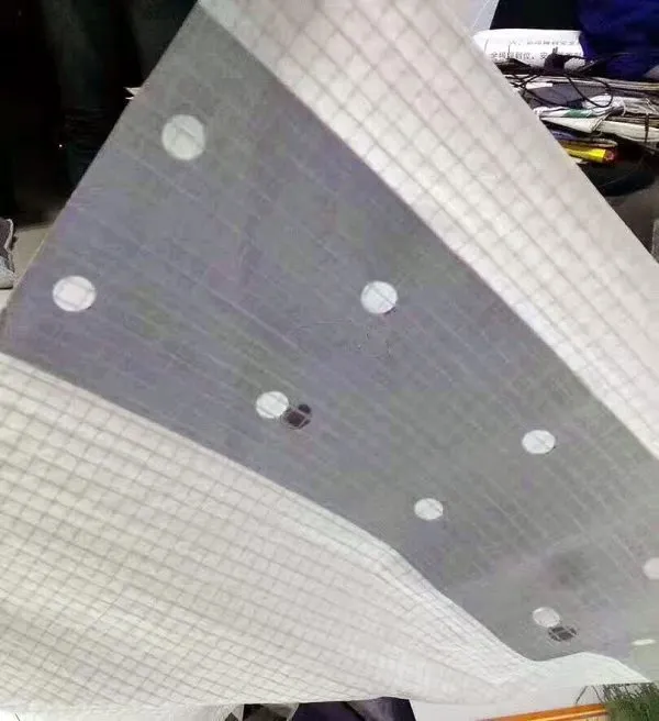 A Clear String Reinforced Poly Tarp – 7 oz FR Scaffold Sheeting with a grid pattern and multiple circular holes is held up at an angle.