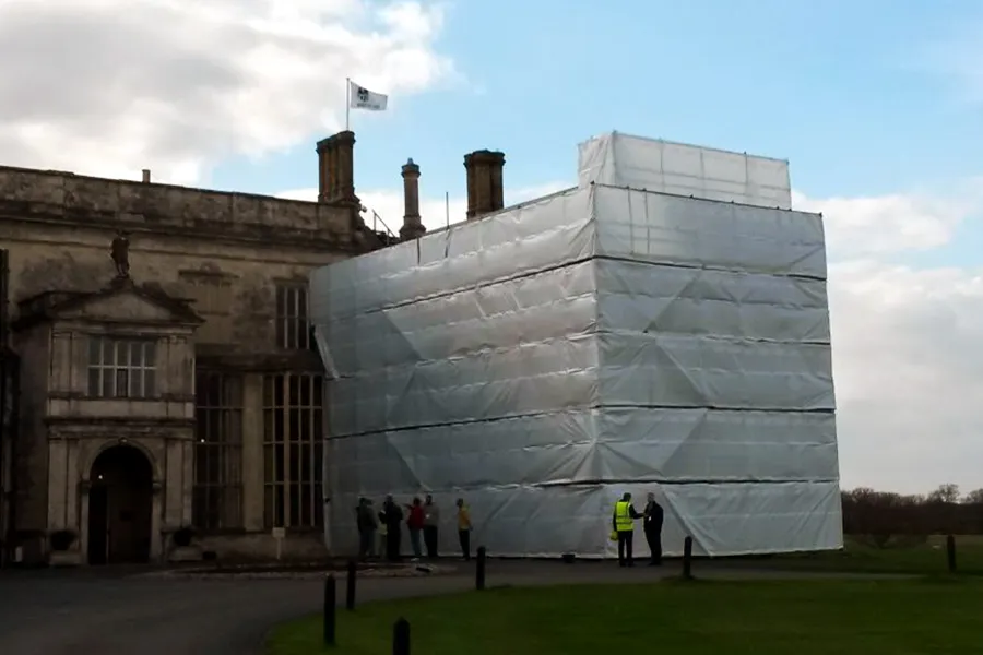 A large section of a historic building is covered in Clear String Reinforced Poly Tarps – 7 oz FR Scaffold Sheeting for renovation work. Several people are standing near the covered area, and a flag is flying atop the building.
