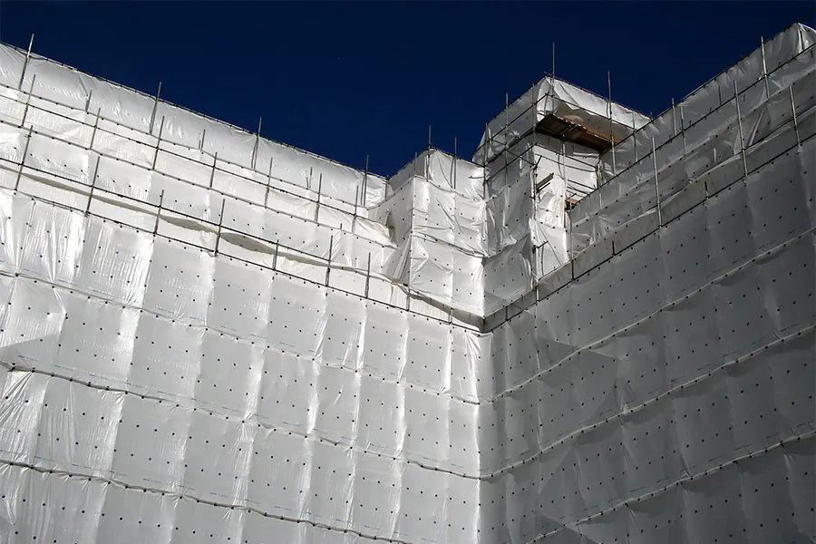 A building under construction is fully covered with Clear String Reinforced Poly Tarps – 7 oz FR Scaffold Sheeting beneath a clear blue sky, with scaffolding visible along the top edges.