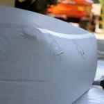 Close-up of a Cube collapsible water tank with round corner reinforced, showcasing its gray exterior and metal D-rings on one side. The background reveals a blurry orange vehicle and some greenery.