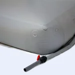 Close-up of the Cube collapsible water tank with round corners reinforced, featuring a hose and red valve attached. The gray inflatable structure rests on a light-colored surface, with multiple metal rings visible on its side.