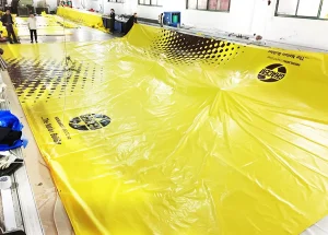 Several people are working with large sheets of Custom Printed Tarps, branded with "HESLY" logos, inside a well-lit industrial space.