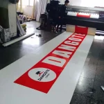 A person operates a large printing machine producing a long banner using Custom Printed Tarps with the word "Dangote" and logo. The banner is predominantly white with large red letters and features a logo in a white circle.