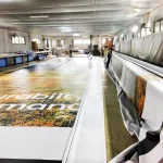 Workers operating large printing machinery in an industrial facility to produce oversized Custom Printed Tarps. The tarps display partially visible text and colorful graphics.