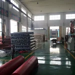A brightly lit factory interior showcasing stacks of Heavy Duty Tarpaulin rolls and machinery. The green floor is marked with yellow lines, while large windows allow natural light to flood the space.