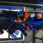 A storage area with Heavy Duty Tarpaulin rolls in various colors, predominantly blue, suspended horizontally from the ceiling near a window with partially closed blue blinds.