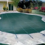 A backyard pool is covered with a Safety swimming pool cover and surrounded by a stone patio, with a house and garden in the background.