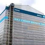 A tall building under construction is covered in scaffolding and FR SCAFFOLD SHEETING-10oz. A large sign on the sheeting reads, "BUILD A BIGGER BUSINESS IMAGE.