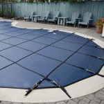 A backyard pool is protected by a Safety swimming pool cover in blue, which is securely fastened with straps and anchors. Adirondack chairs and a potted plant adorn the area along the back fence.