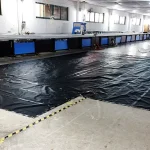A Lumber Tarp with Flap for Flatbed Truck and Trailer – 8′ Drop covers part of the floor in a spacious industrial room with several worktables, windows, and equipment. A few individuals are present in the background.