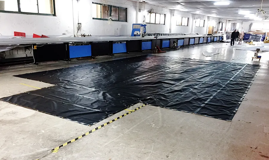 A Lumber Tarp with Flap for Flatbed Truck and Trailer – 8′ Drop covers part of the floor in a spacious industrial room with several worktables, windows, and equipment. A few individuals are present in the background.