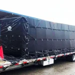 A large rectangular object wrapped in a Lumber Tarp with Flap for Flatbed Truck and Trailer – 8′ Drop is secured on a flatbed trailer parked outside a warehouse.