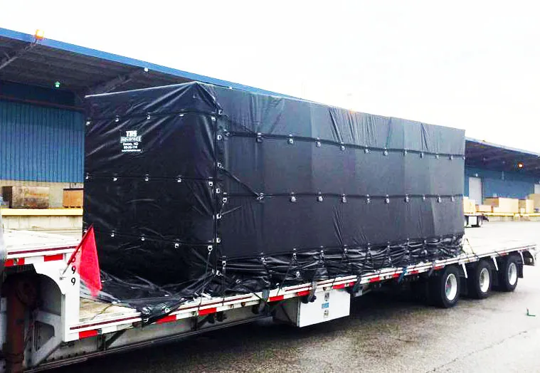 A large rectangular object wrapped in a Lumber Tarp with Flap for Flatbed Truck and Trailer – 8′ Drop is secured on a flatbed trailer parked outside a warehouse.