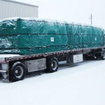 A flatbed truck carrying a large, tarped load is parked on a snowy industrial property. The cargo is covered by the **Lumber Tarp with Flap for Flatbed Truck and Trailer – 8′ Drop**, and there is snow accumulation on the truck and ground.