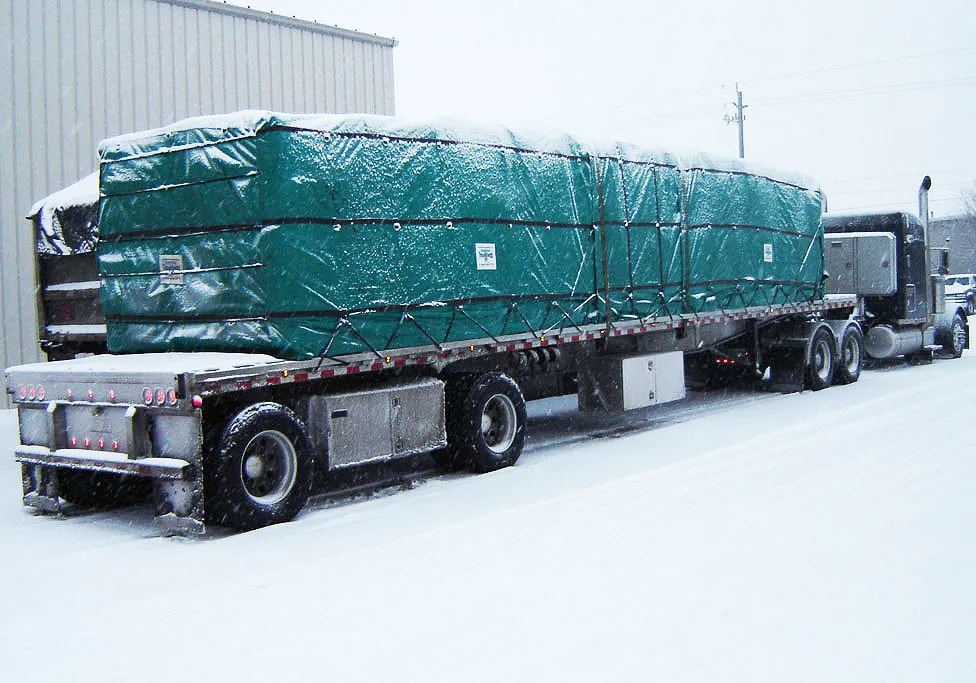 A flatbed truck carrying a large, tarped load is parked on a snowy industrial property. The cargo is covered by the **Lumber Tarp with Flap for Flatbed Truck and Trailer – 8′ Drop**, and there is snow accumulation on the truck and ground.