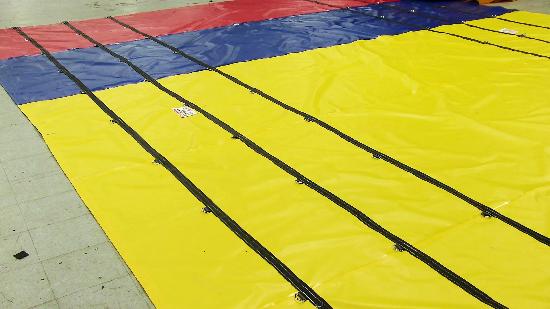 Close-up view of a large, colorful Lumber Tarp with Flap for Flatbed Truck and Trailer – 8′ Drop, divided into sections of yellow, blue, and red, with black straps laid across its surface.