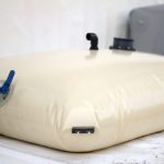 A Pillow water tank with metal pads in corners, featuring an attached hose and valves, placed on a white surface.