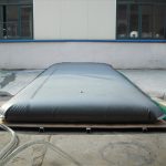 A large Pillow water tank with metal pads in the corners is laid out on a wooden platform outdoors, with hoses connected to it. Three large windows are visible in the background.