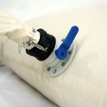 A close-up view of the Pillow water tank with metal pads in the corners, featuring a blue lever valve attached to a nozzle and secured with a metal hose clamp.