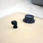 Close-up of a Pillow water tank featuring metal pads in the corners, with a round cap and an elbow pipe fitting on its flat surface. The background is blurred and appears to be a light-colored surface.