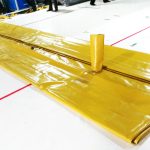 A large yellow pillow water tank with metal pads in the corners lies flat on a white floor, with two upright cylindrical openings visible.