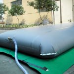 A large gray Pillow water tank with metal pads in the corners and a connected hose is placed on a green mat outdoors near a building.