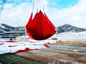 A crane suspends a large red container equipped with Snow Tarps over a snowy, industrial area with mountains in the background.